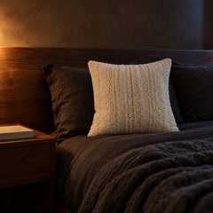 Ivory woven cushion on a dark linen bed at night